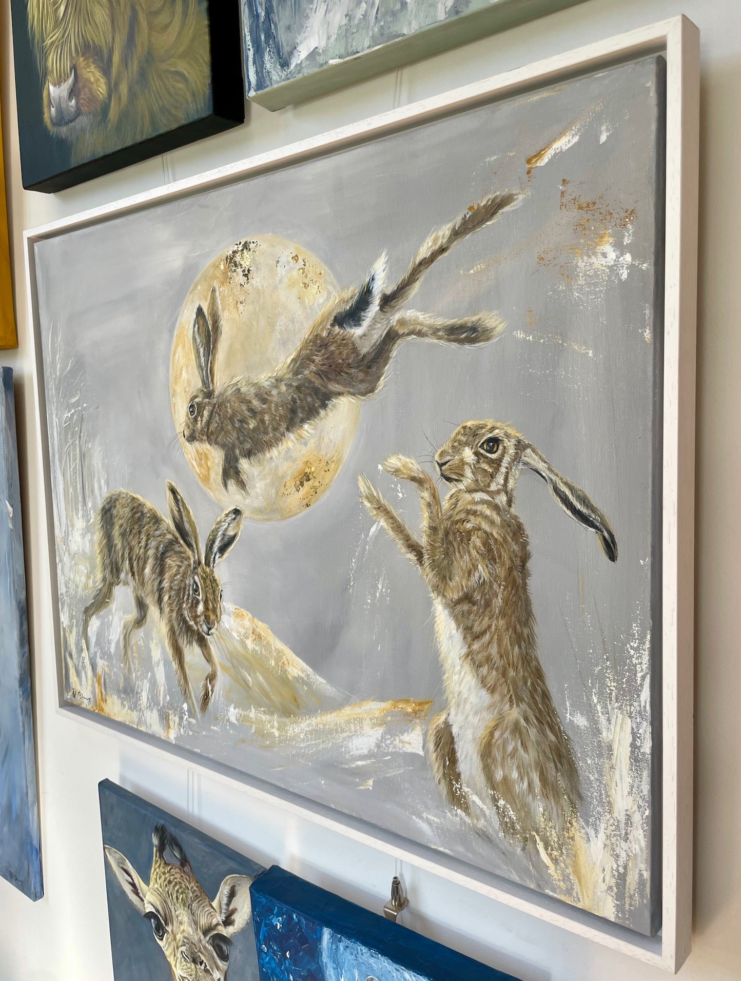 Side view of painting of three hares playing in the snow and jumping over the moon in white frame by Debbie Storey